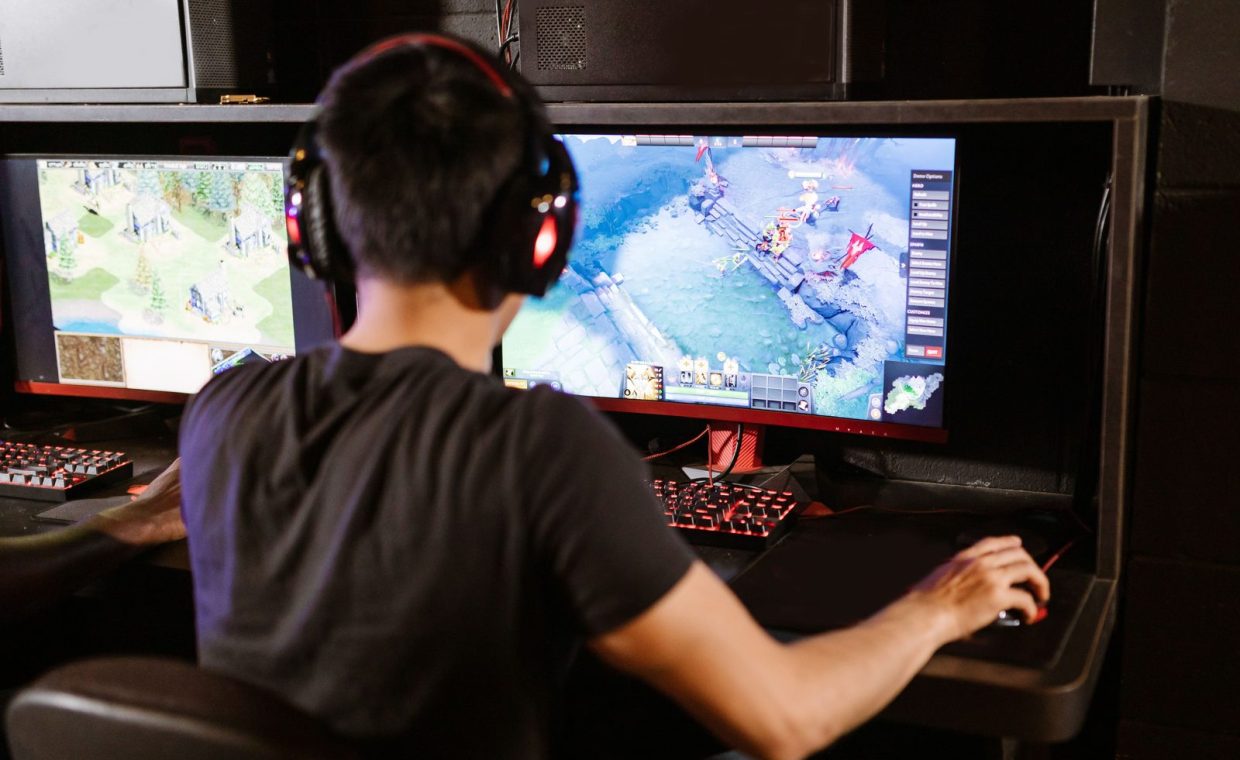 Adult male gamer immersed in PC gaming on dual monitors with headphones indoors.