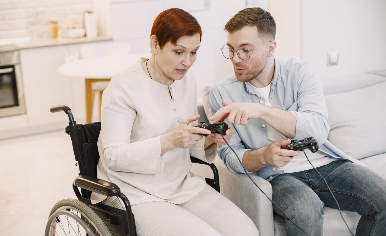 Older woman in wheelchair and young man play video games together, fostering connection.
