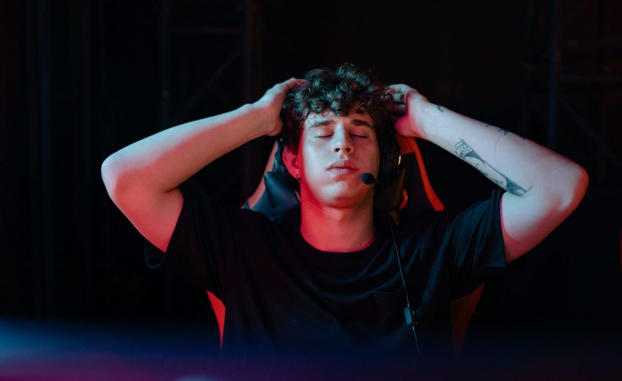 Young man with headset and tattoos, taking a break from gaming with eyes closed, seated indoors.