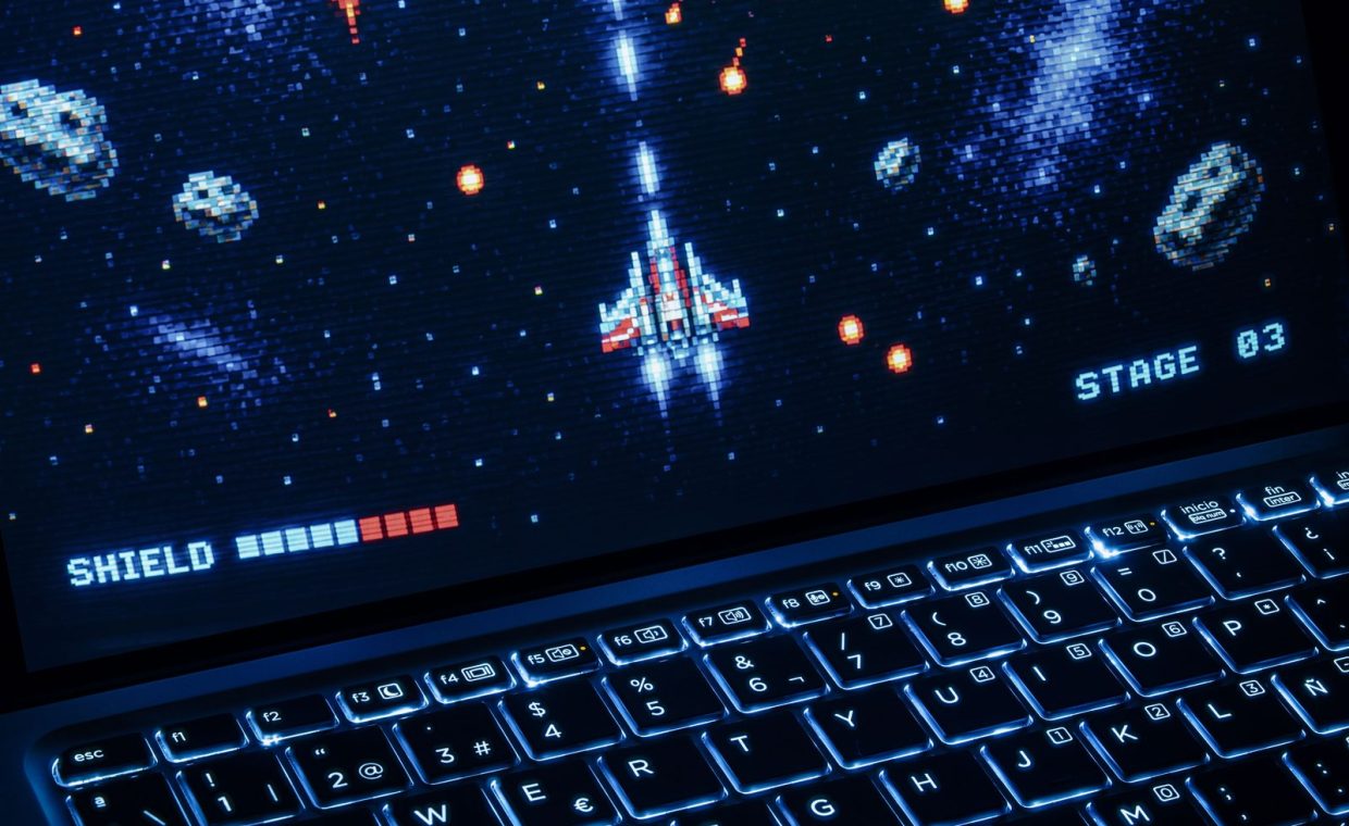 A classic arcade space shooter game displayed on a laptop with illuminated keyboard, showing retro graphics.