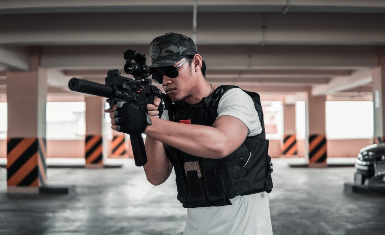 A man dressed in tactical gear aims a rifle in a shaded parking area, suggesting a training scenario.