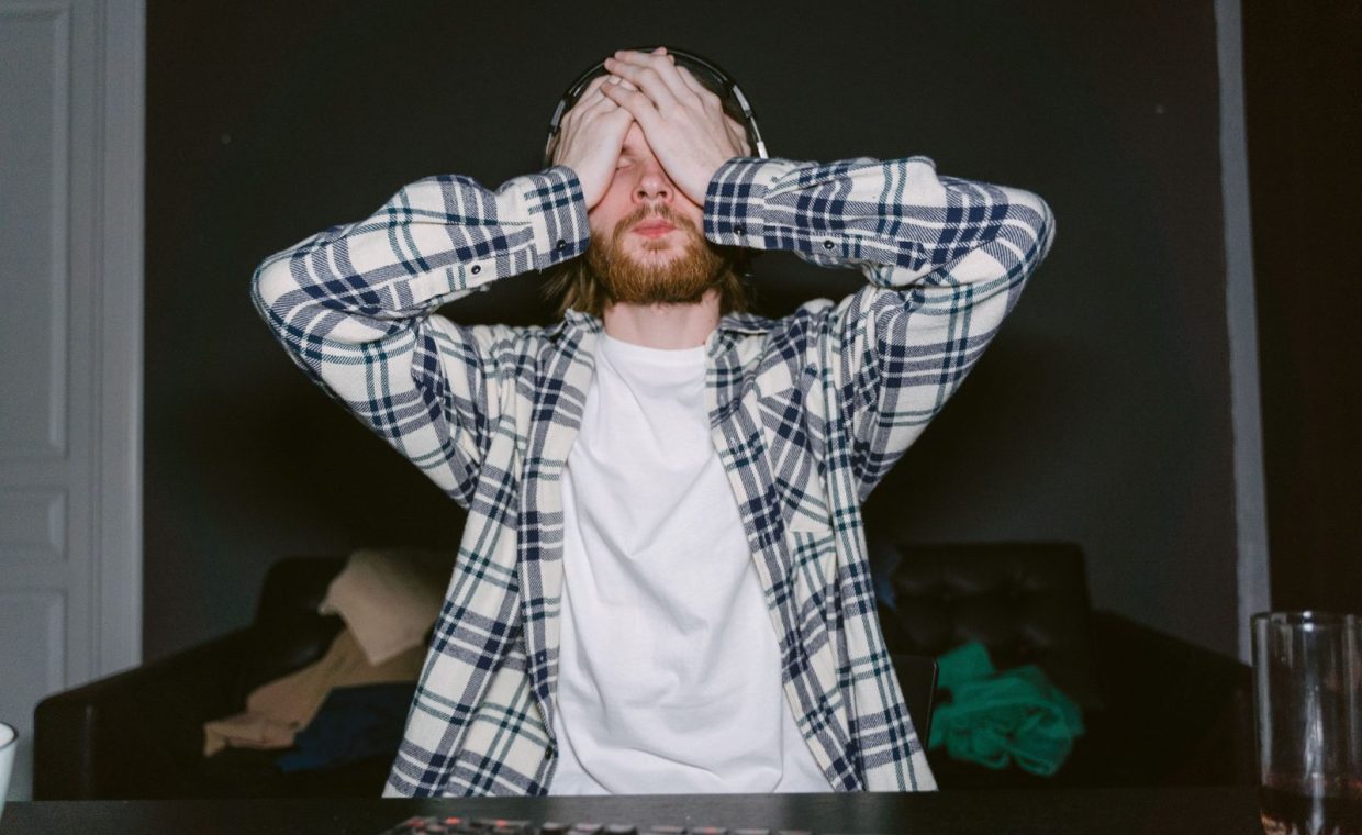 Focused male gamer in plaid shirt, showing stress with hands on face, indoors.
