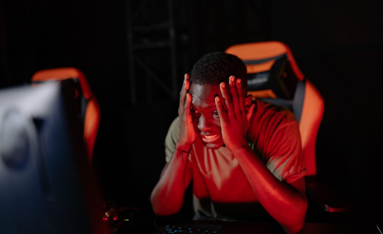 A man intensely reacts to a game loss at his computer in a dimly lit gaming room.