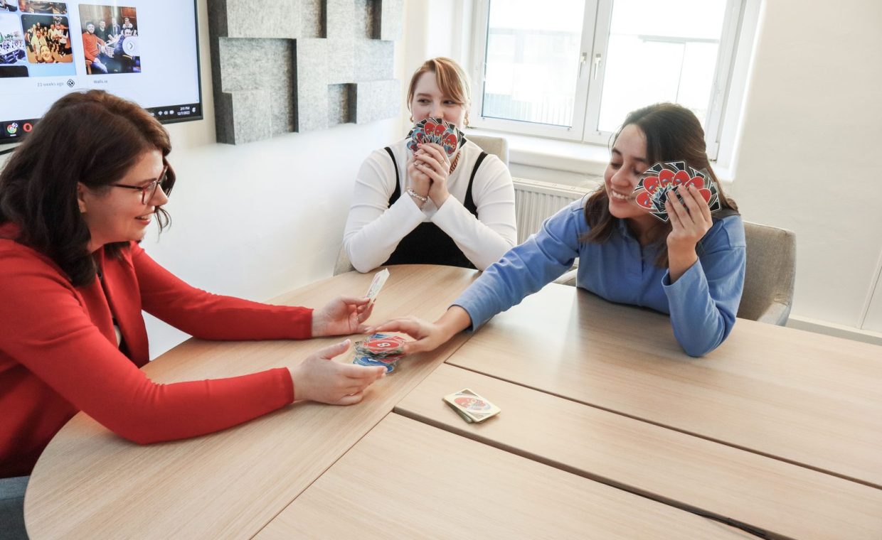 Three women playing cards in a modern office, enhancing team bonding.