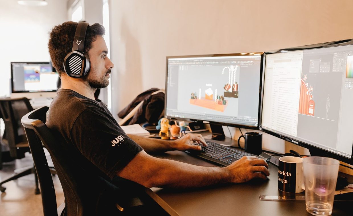 A man wearing headphones works on a computer with two monitors in a modern office setup.