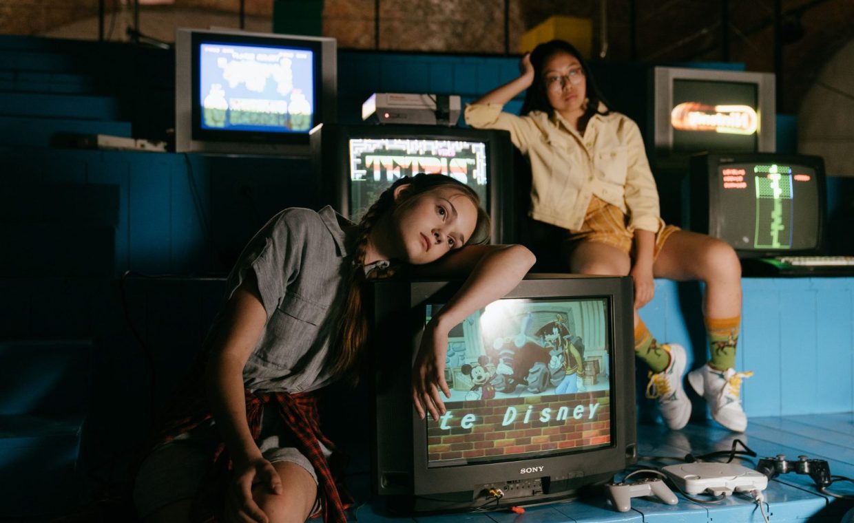 Two women in casual outfits immersed in retro gaming with vintage televisions and game consoles.